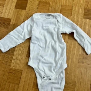 Vit långärmad body från Hello Baby - En basic vit långärmad body från Hello Baby i mjuk bomull. Klassisk modell med rund hals och tryckknappar i grenen för enkel påklädning. Perfekt att ha under andra plagg eller som den är.