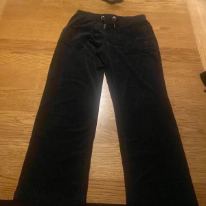 Svarta mjukisbyxor Juicy Couture - Svarta mjukisbyxor från Juicy Couture med breda ben och snörning i midjan. Byxorna har fickor både fram och bak med snygga detaljer och är tillverkade i mjuk sammet som ger en lyxig känsla. Perfekta för en chill och trendig look.
