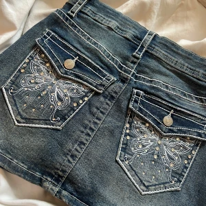 Jeanskjol med broderade fickor  - Snygg jeanskjol i tvättad blå denim. Kjolen har coola broderade bakfickor med fjärilsmotiv och silvriga nitar, samt klassiska jeansdetaljer som bälteshällor och knappar. Perfekt för dig som vill ha en unik och trendig look.