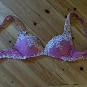 Rosa spets-bh med blommig spets - Säljer en söt rosa bh med vit blommig spetsdetalj över kuporna och axelbanden. Bh:n har vadderade kupor och justerbara axelband. Perfekt för dig som gillar romantiska och feminina underkläder.