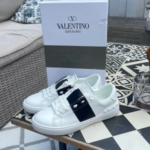 Valentino Garavani Open sneakers vit/svart - Snygga Valentino Garavani Open sneakers i vitt skinn med bred svart läderdetalj över mitten. Klassisk rund tå, vita snören och ikoniska nitar på hälen. Perfekta för dig som gillar lyxiga och stilrena sneakers med en twist. Dom är helt oanvända och allting följer med 