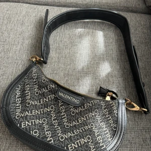 Valentino väska - Snygg svart axelväska från Valentino med logomönster över hela väskan.Den enda nackdelen är att låset fastnar lite, därav priset, men väskan är i mycket bra skick.