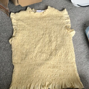 Gul volangtopp med smock - Supersöt ljusgul topp med smockad framsida och volangdetaljer vid ärmhål, hals och nederkant. Ärmlös modell i mjuk bomull, perfekt för varma dagar. Enkel att styla med jeans eller kjol för en fräsch look.