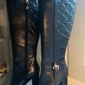 Svarta höga stövlar Louis Vuitton - Snygga svarta höga stövlar från Louis Vuitton i läder med präglad monogramdetalj upptill, silverfärgade nitar och spänne vid sidan. Spetsig tå och hög klack ger en edgy vibe. Dragkedja på insidan för enkel påtagning.