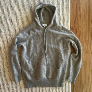 Davida Kashmir hoodie  - Mjuk och lyxig grå hoodie i kashmir från Davida. Tröjan har huva, dragsko och fickor framtill. Perfekt för en avslappnad och stilren look. Klassisk passform och ribbade muddar vid ärmslut och nederkant. Storlek är M men den är tvättad så storlek blir XS. Sydd vid armhåla men syns ej.