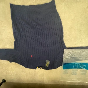 Mörkblå stickad tröja Polo Ralph Lauren - Helt ny Mörkblå kabelstickad tröja från Polo Ralph Lauren med klassisk röd broderad logga på bröstet. Tröjan har rund halsringning och långa ärmar, perfekt för dig som gillar en stilren och tidlös look. Materialet är mjukt och skönt, passar till många outfits.