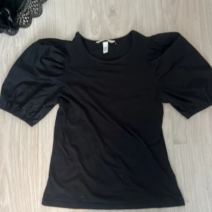 Svart topp med puffärmar från H&M - Svart topp från H&M i storlek XS med trendiga puffärmar och rund halsringning. T-shirten har en enkel, stilren design och är tillverkad i mjukt bomullsmaterial. Perfekt för dig som gillar en klassisk look med en twist.