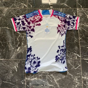 Spanien Adidas fotbollströja S - Säljer en officiell Spanien fotbollströja från Adidas i storlek S. Tröjan är vit med blå och lila detaljer, har korta ärmar och ett unikt mönster på axlar och ärmar. Tre blå ränder på axlarna och rosa detaljer vid ärmslut och krage. Tillverkad i lätt och ventilerande polyester.