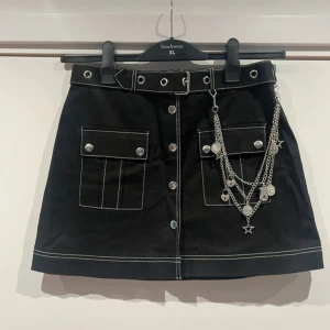 Limited edition svart kortkjol Dolls Kill x Bratz - Limited edition slut såld svart kortkjol med vita kontrastsömmar från Dolls Kill x Bratz collab.  Kjolen har två stora fickor framtill, bälte med metallöljetter och coola silverkedjor med stjärn- och myntdetaljer. Stängs med knappar framtill. Materialet är bomull med lite stretch. Importerad från usa. Collectible item. 