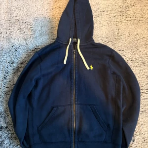 Marinblå hoodie från Polo Ralph Lauren - Säljer en marinblå hoodie från Polo Ralph Lauren med dragkedja och huva. Tröjan har vita snören, två fickor framtill och den klassiska gula loggan broderad på bröstet. Perfekt för dig som gillar stilrena och bekväma plagg.