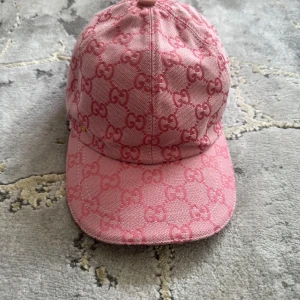 Rosa Gucci keps med monogram - Snygg rosa keps från Gucci med klassiskt GG-monogram över hela kepsen. Kepsen har böjd skärm och är tillverkad i ett slitstarkt bomullstyg. Perfekt accessoar för att lyfta din outfit med en lyxig touch.   STORLEK L (59 cm)