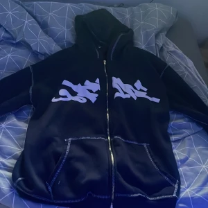 Svart hoodiejacka med tryck - Svart hoodiejacka med dragkedja och stora vita tryck på bröstet. Jackan har huva, synliga sömmar och fickor framtill. Perfekt för en streetwear-look och chill dagar.