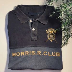 Morris R. Club pikétröja i mörkblått - Snygg mörkblå pikétröja från Morris R. Club med gul brodyr och tryck över bröstet. Klassisk krage, två knappar och emblem på vänster sida. Tröjan är i bomull och har en sportig, stilren vibe som passar perfekt till jeans eller chinos.