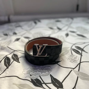 Louis Vuitton Bälte - Snyggt Louis Vuitton-bälte i svart och grått med rutigt mönster och ikoniskt LV-spänne i silver. Bältet är gjort i skinn med en brun insida och har tydlig Louis Vuitton Paris-stämpel. Perfekt accessoar för att lyfta din outfit.