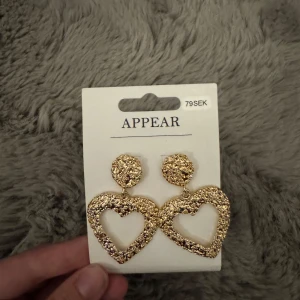 Guldiga hjärtformade örhängen Appear - Säljer ett par statement örhängen från Appear i guldig färg. De har en cool, texturerad yta och är formade som stora hjärtan som hänger från runda plattor. 