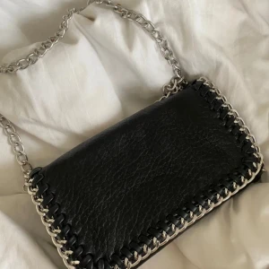 Svart axelväska med kedja - Snygg svart axelväska i skinn med grov silverfärgad kedja som axelrem. Väskan har coola detaljer med flätad kant runtom och en stilren, rektangulär form. Perfekt för dig som vill ha en edgy accessoar till din outfit.