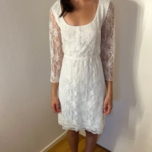 Vit spetsklänning med trekvartsärm - Superfin vit klänning i spets med trekvartsärmar och rundad urringning. Klänningen är kort och har ett vackert blommigt spetsmönster över hela plagget.