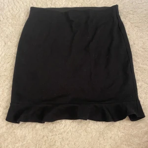 Svart kortkjol med volangkant - Enkel och snygg svart kortkjol med volangdetalj längst ner. Kjolen har en rak passform och är perfekt för dig som gillar stilrena plagg med en liten twist. Passar till många olika outfits och är lätt att matcha.