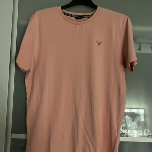 Ljusrosa t-shirt från GANT - Snygg ljusrosa t-shirt från GANT med liten broderad logga på bröstet. Klassisk rund halsringning och korta ärmar. Tillverkad i mjuk bomull som känns skön mot huden. Perfekt basic-plagg med clean vibe.