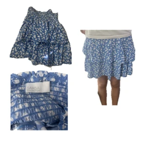 Blommig blå volangkjol Samsa Collection - Supersöt blå kortkjol med vita blommor från Samsa Collection. Kjolen har volanger och resår i midjan för en bekväm passform. Perfekt för dig som gillar en feminin och lekfull stil. Lätt och luftig, passar perfekt till varma dagar.