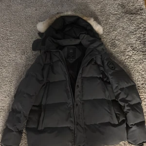 Canada Goose Black Label - Säljer en svart pufferjacka från Canada Goose med stor huva och snygg pälsdetalj. Jackan har klassisk patch på ärmen, dragkedja och knappar framtill. Perfekt för kalla vinterdagar och riktigt stilren look.