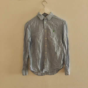 Polo Ralph lauren skjorta - Snygg ljusblå skjorta från Polo Ralph Lauren i relaxed fit. Tillverkad i linne med klassisk krage, knäppning framtill och lång ärm. Ikoniska gröna broderade loggan på bröstet ger en clean och stilren vibe.