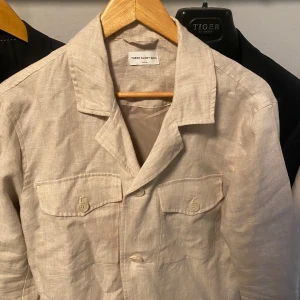Beige linnekavaj från These Glory Days - Snygg beige overshirt i linne från These Glory Days. Klassisk krage, två bröstfickor med knappar och enkel knäppning framtill. Perfekt för dig som gillar en clean och stilren look med avslappnad känsla.