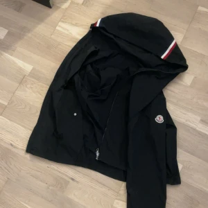 Svart vindjacka från Moncler med huva - Svart vindjacka från Moncler storlek xs kan passa s sprillans ny och med huva och dragkedja. Jackan har Moncler-logga på ärmen och en snygg detalj med röd, vit och blå rand på huvan. Perfekt för dig som vill ha en stilren och sportig look 