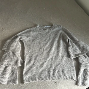 Grå stickad volangtröja från NA-KD - Superfin ljusgrå stickad tröja från NA-KD med unika volangärmar i tre lager. Tröjan har rund halsringning och är ribbstickad vid ärmslut och nederkant. Perfekt för dig som vill ha något extra till din outfit.