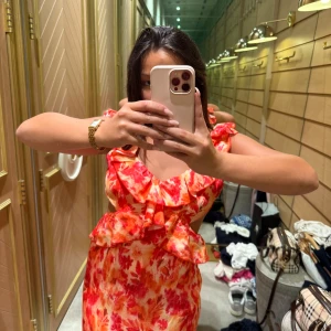 Blommig volangklänning med öppen rygg - Superfin långklänning med blommigt mönster i rött, orange och vitt. Klänningen har volangdetaljer vid axlar och byst, öppen rygg med knyt och är ärmlös. Perfekt för sommarens alla tillfällen när du vill sticka ut lite extra.