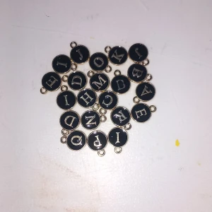 Svarta bokstavsberlocker i metall - Säljer flera runda svarta berlocker med bokstäver i silverfärg och guldkant. Perfekta att använda till armband, halsband eller örhängen för att skapa personliga smycken. Varje berlock har en ögla för enkel fastsättning.