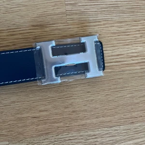 Mörkblått H- skärp med H-spänne - Snyggt bälte med ikoniskt H-spänne i silverfärgad metall. Mörkblått med vit söm. Bältet är gjort i exklusivt skinn och passar perfekt till jeans eller kostymbyxor. Oanvänd men har ej prislapp.