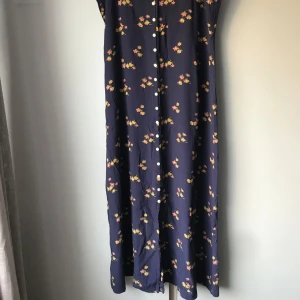 Marinblå blommig långklänning - Snygg  långklänning med små blommor i gult och rosa. Klänningen har knappar hela vägen framtill och är ärmlös, perfekt för varma dagar. Lätt och luftig modell som ger en avslappnad men ändå stilren look.