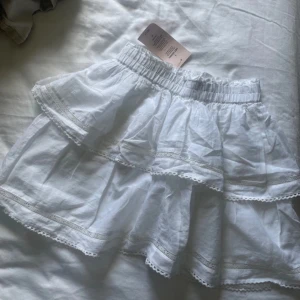 Vit volangkjol med spets från Nelly - Supersöt vit kortkjol från Nelly med volanger och fina spetsdetaljer. Kjolen har resår i midjan och är gjord i bomull, vilket gör den både luftig och bekväm. Perfekt för sommarens alla tillfällen när du vill känna dig extra söt. Aldrig använd. 