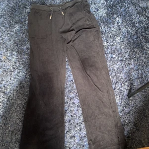 Svarta mjukisbyxor från Juicy Couture - Svarta mjukisbyxor från Juicy Couture med broderad JC-logga framtill. Byxorna har dragsko i midjan, raka ben och mjukt, sammetsliknande material. Fickor både fram och bak med snygga detaljer. Perfekta för en chill och trendig look.