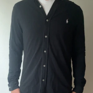Svart skjorta från Ralph Lauren - Svart långärmad skjorta från Ralph Lauren i lätt mesh-bomull. Klassisk design med knappar framtill och vit broderad logga på bröstet. Skjortan har en normal passform och är perfekt för dig som gillar stilrena plagg med premiumkänsla.