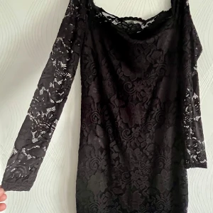 Svart spetsklänning från AMISU - Supersnygg svart kortklänning från AMISU i genombruten spets med blommigt mönster. Klänningen har långa ärmar och en feminin, figurnära passform. Perfekt för dig som vill sticka ut med en elegant och trendig look.