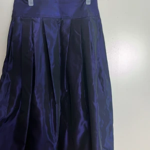 Mörklila långkjol från Astrid Bachmann - Snygg mörklila långkjol i glansigt tyg från Astrid Bachmann. Kjolen har bred linning, djupa veck och dragkedja på sidan. Perfekt för dig som vill sticka ut med en elegant och klassisk look. Okänd storlek men måtten är 78 på längden, och 78 runt midjan. 🥰