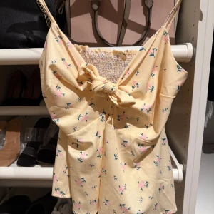 Gul blommig linnetopp med knyt - Supersöt ljusgul linnetopp med små rosa blommor och gröna blad. Toppen har smala axelband, knyt framtill och smockad rygg för extra komfort. Perfekt för varma dagar och ger en fräsch sommarkänsla till din outfit.
