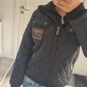 Svart quiltad jacka Geographical Norway - Quiltad svart jacka från Geographical Norway med coola detaljer och orange patch på bröstet. Jackan har dragkedja framtill, ribbad krage och långa ärmar. Perfekt för dig som gillar en sportig och urban stil. Passar höst och vinter.