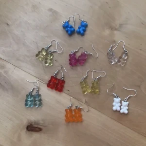 Färgglada örhängen med nallebjörnar - Säljer flera par örhängen med söta nallebjörnsberlocker i olika färger: blå, röd, rosa, vit, gul, orange, grön och transparent. Hängena är i plast och har krokar i metall. Perfekta för dig som vill ha en lekfull och unik stil. Det finns ett par av alla färger i lager förutom de transparenta (2 par) och de ljus gula (3 par), (som är brevid de rosa)💞Man kan även köpa alla 12 par för 40kr💞