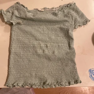 Ljusgrön smockad topp med volang - Supersöt ljusgrön smockad topp med korta ärmar och volangdetaljer vid ärmslut och hals. Toppen är stretchig och har en ribbad struktur som ger en snygg passform. Är öppen för bud