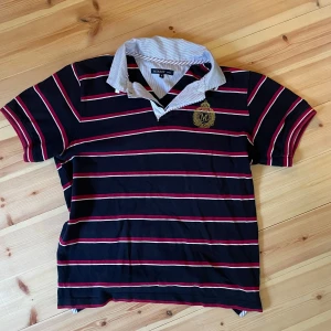 Randig pikétröja från Murrey - Snygg pikétröja från Murrey i marinblått med röda och vita ränder. Tröjan har klassisk krage med tunna ränder och ett broderat emblem på bröstet. Perfekt för en preppy look och passar till jeans eller shorts.