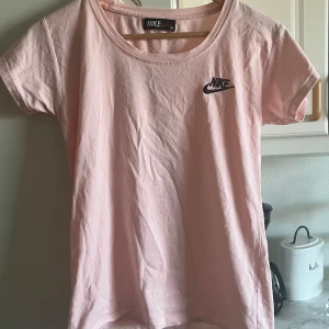 Ljusrosa Nike t-shirt i bomull - Snygg ljusrosa t-shirt från Nike med klassisk logga broderad på bröstet. T-shirten har rund halsringning och korta ärmar. Tillverkad i mjuk bomull som är skön att bära hela dagen. Perfekt för en sportig och avslappnad stil. Toppen är endast använd 1 gång! 