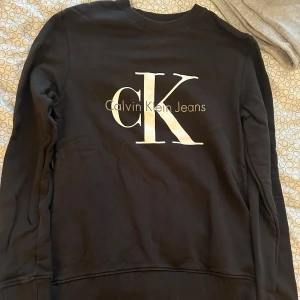 Svart Calvin Klein Jeans sweatshirt XS - Svart sweatshirt från Calvin Klein Jeans i storlek XS. Tröjan har en stor vit CK-logga framtill och rund halsringning. Tillverkad i mjuk bomullsmix med ribbade muddar vid ärmslut och nederkant. Perfekt för en clean och stilren look.