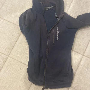 Marinblå fleeceväst Sail Racing - Snygg marinblå fleeceväst från Sail Racing med hel dragkedja och huva. Västen har en bröstficka med dragkedja och logga på bröstet. Perfekt för lager-på-lager och chill dagar.