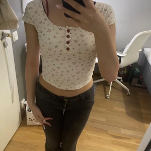 Blommig croppad topp med blommor  - Supersöt croppad topp i ljusbeige med små rosa blommor och röda knappar framtill. Toppen har korta ärmar, rundad halsringning och figurnära passform. Röda detaljer vid axeln, fram och bak. Perfekt för dig som gillar feminin stil. Prislappen är klippt men har aldrig använt den!