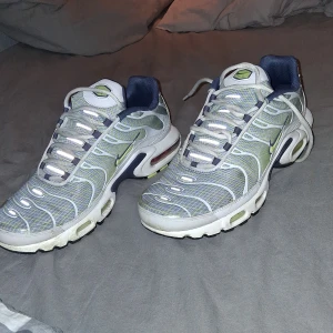 Nike Air Max Plus Tn sneakers vit/blå - Nike Air Max Plus Tn sneakers i vitt och blått med limegröna detaljer. Skorna har mesh-överdel, synliga Air Max-enheter i sulan och Tn-logga bak. Snygg vågig design och snörning framtill. Perfekta för dig som gillar streetwear och unika sneakers.
