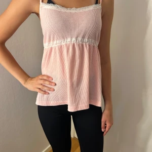 Ljusrosa linnetopp med spetsdetaljer - Supersöt ljusrosa linnetopp med tunna axelband och vit spets längs urringningen och under bysten. Toppen har en luftig och flowy passform, perfekt för varma dagar. Materialet på linnet är ribbat.