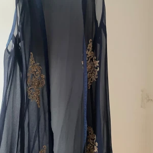 Mörkblå långklänning med guldiga broderier - Säljer en elegant mörkblå långklänning i transparent tyg med vackra guldiga broderier längs sidorna. Klänningen har en flowy passform och ger en lyxig känsla med sina detaljer. Perfekt för dig som vill sticka ut med stil.